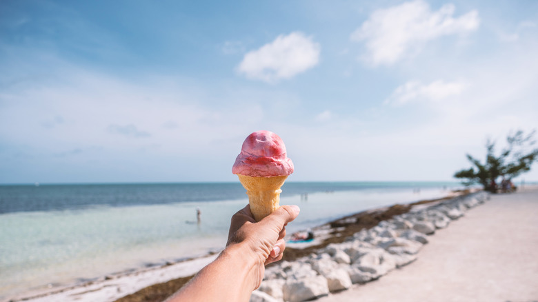 Hand holding an ice cream cone against a beach scene