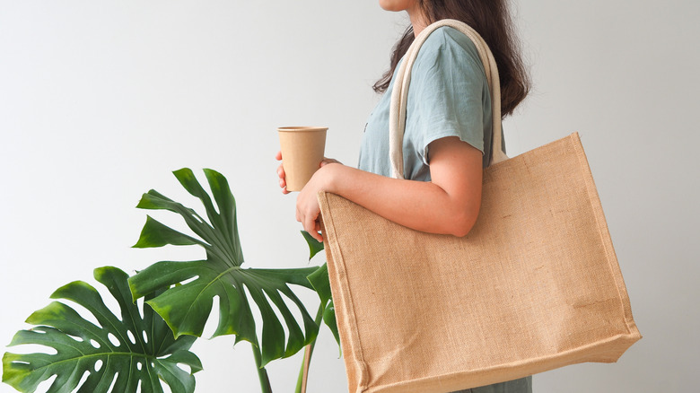 A woman holding a brown paper coffee cup and wearing a jute reusable shopping bag next to a potted monstera plant