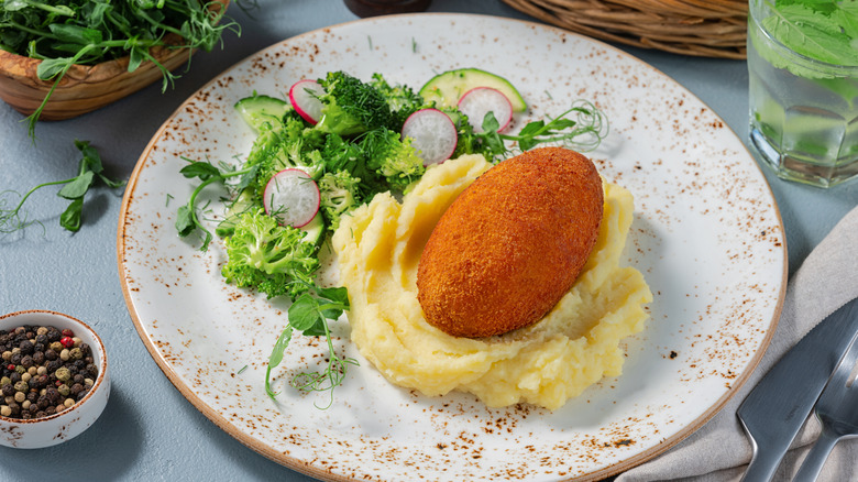 Chicken Kiev sits on a plate on top of mashed potatoes, with broccoli and radishes on the side.