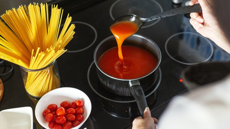 A home cook stands over a pot of tomato sauce on the stove, next to a jar of uncooked spaghetti and a bowl of cherry tomatoes