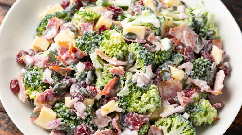 Top down view of a broccoli salad in a white bowl featuring visible cranberries, pumpkin seeds, cheese, bacon, and broccoli in a creamy dressing