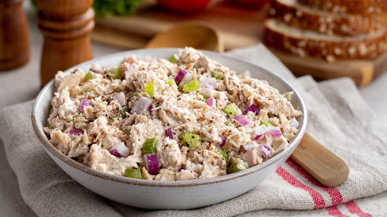 A white bowl of tuna salad with celery and red onion, on a table next to a spoon and bread