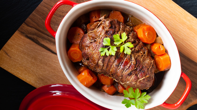 Top view of a pot roast in a enamel pot surrounded by carrots, sitting on a wooden cutting board