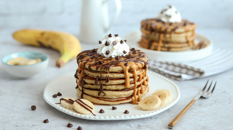 A stack of banana peanut butter pancakes topped with a dollop of cream, chocolate chips, chocolate sauce and peanut butter
