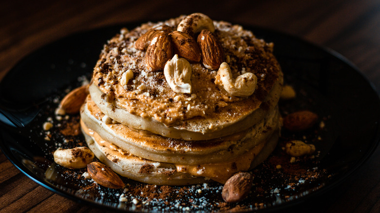 A stack of peanut butter pancakes covered in peanut butter and topped with almonds and cashews