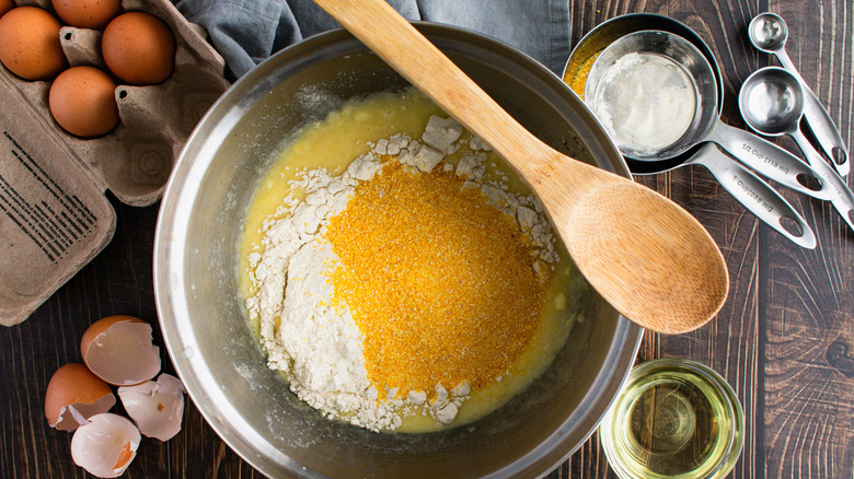 a mixing bowl full of cornbread ingredients with a wooden spoon resting on the rim and additional ingredients and measuring cups beside it.