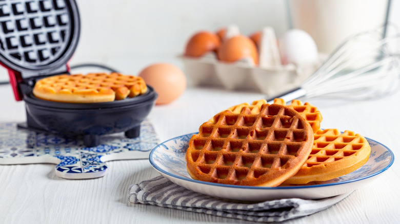 Waffles freshly cooked on a plate with waffles cooking in a waffle iron behind it