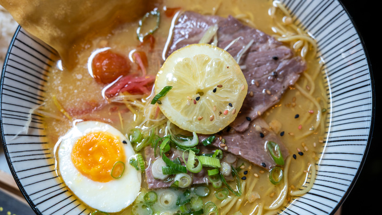 A bowl of ramen featuring thin sliced beef, scallions, half of a hard boiled egg, and noodles in broth with a slice of lemon on top