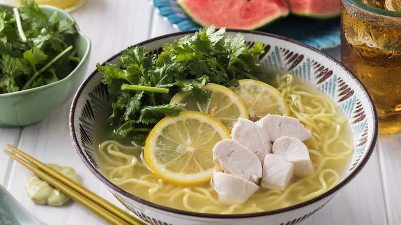 A bowl of ramen with chopped chicken, cilantro, and lemon slices on top. There is more cilantro off to the side as well as a pair of chopsticks, a drink, and some watermelon in the background