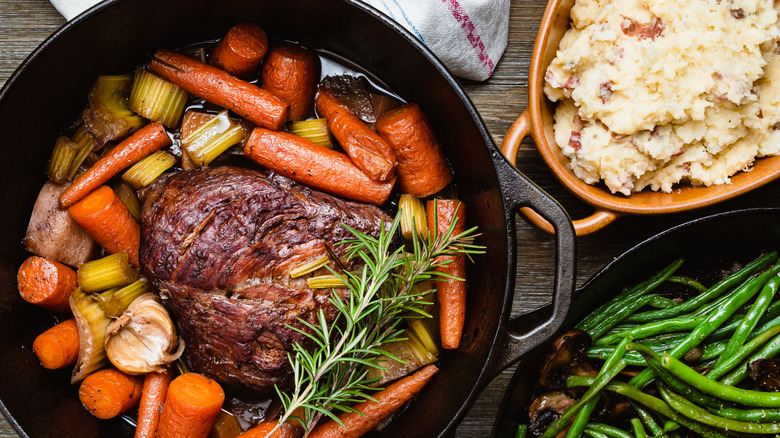 A beef roast and vegetables in cast iron skillet alongside green beans and mashed potatoes