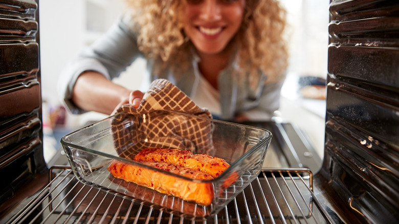 woman pulls out baked salmon in glass dish from oven