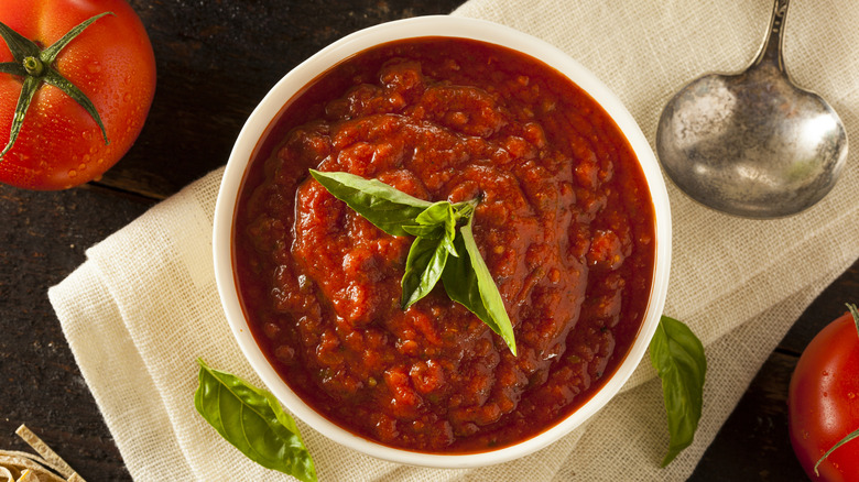 A bowl of marinara sauce topped with fresh basil sitting on a white napkin with fresh tomatoes and garlic nearby