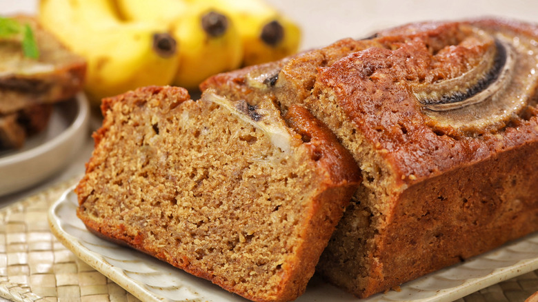 A loaf of banana bread on a platter, with one slice resting against the rest of the loaf, and several bananas visible in the background