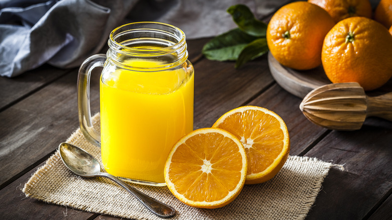 A mason jar glass of orange juice rests on a mat on a wooden table, next to a spoon and a sliced orange, with several whole oranges piled together in the background