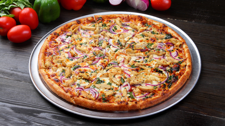 A chicken pizza with onions and tomatoes is served on a pizza tray with tomatoes on a table.