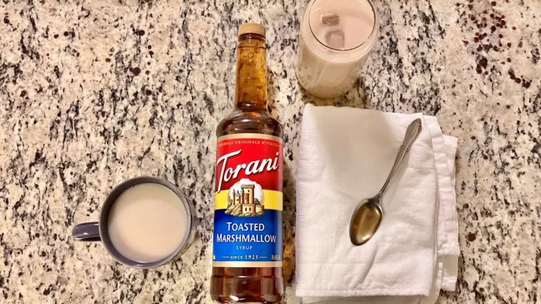 A bottle of Torani Toasted Marshmallow Syrup sits on a marbled surface beside a cup of coffee, a glass of iced coffee, and a teaspoon resting on a kitchen-towel
