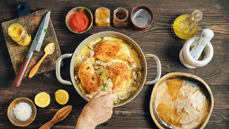 A person cooking a creamy chicken skillet on a wooden table, with numerous ingredients and utensils nearby