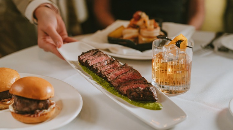 a hand setting down a plate of sliced steak next to a cocktail on a white tablecloth