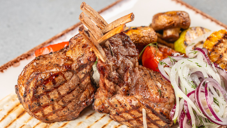 Grilled lamb chops, one with two ribs and one with three, on a plate with some dressed onions, tomato, and mushrooms