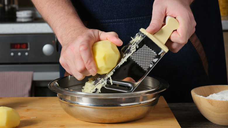 Someone grating a peeled potato on an aluminum box grater