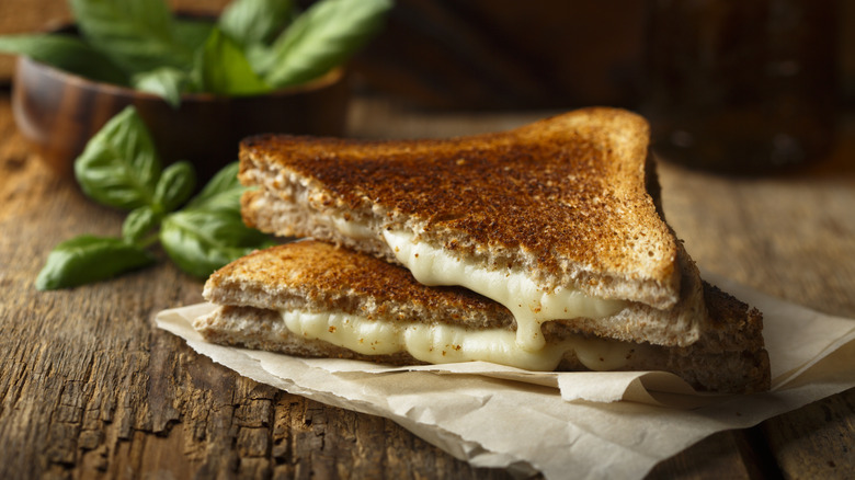 A grilled cheese sandwich sliced in half and oozing with white cheddar, in front of a bowl of basil