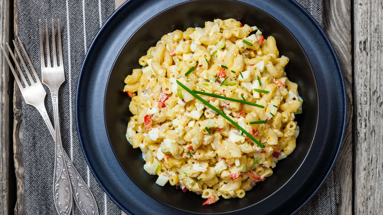A large portion of Southern macaroni salad in a black bowl, set on a matching plate, resting on a wooden table next to two forks