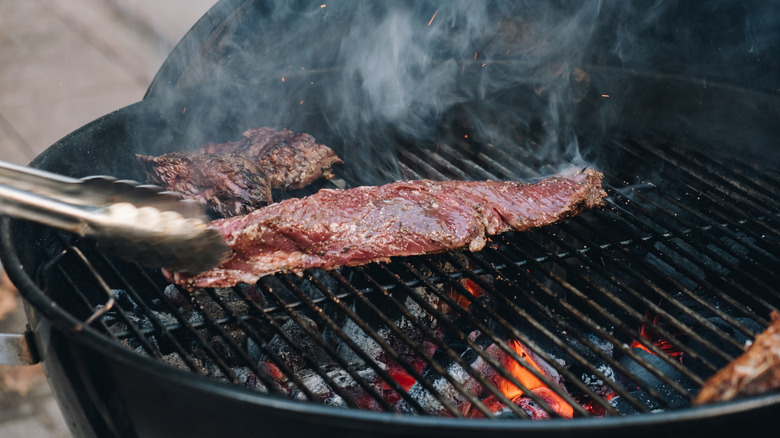 A pair of tongs grasp a piece of skirt steak on a charcoal grill, smoke rising from the meat.