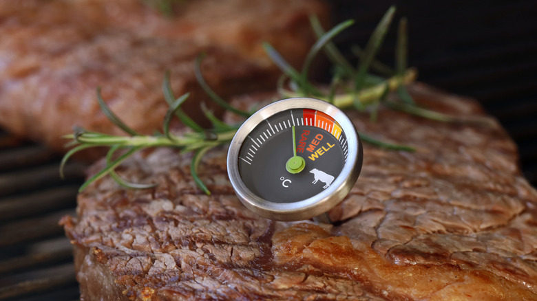 A thermometer sticking out of a piece of cooked beef on a grill. The temperature is just below rare.