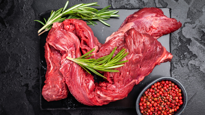 Two pieces of skirt steak folded over on a black cutting board with rosemary and other seasonings.