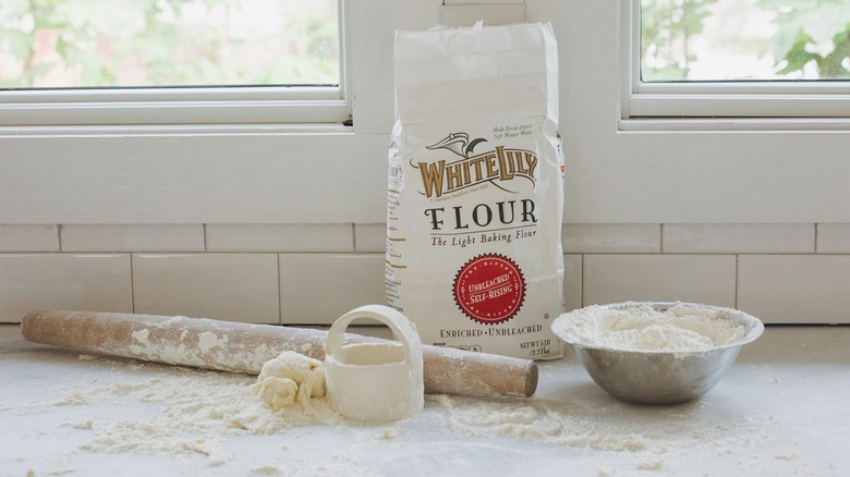 Bag of White Lily self-rising flour on a counter beside a rolling pin, a metal bowl, and dough