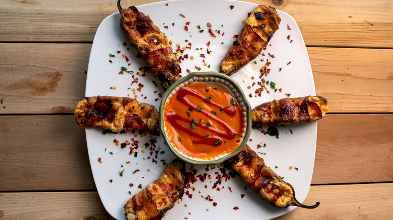 A plate of bacon wrapped jalapeño poppers with a bowl of spicy sauce in the middle