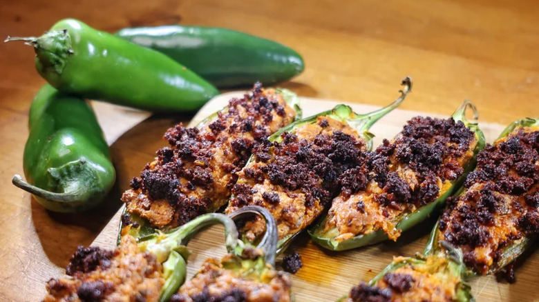A cutting board filled with chorizo topped jalapeño poppers next to three fresh jalapeños
