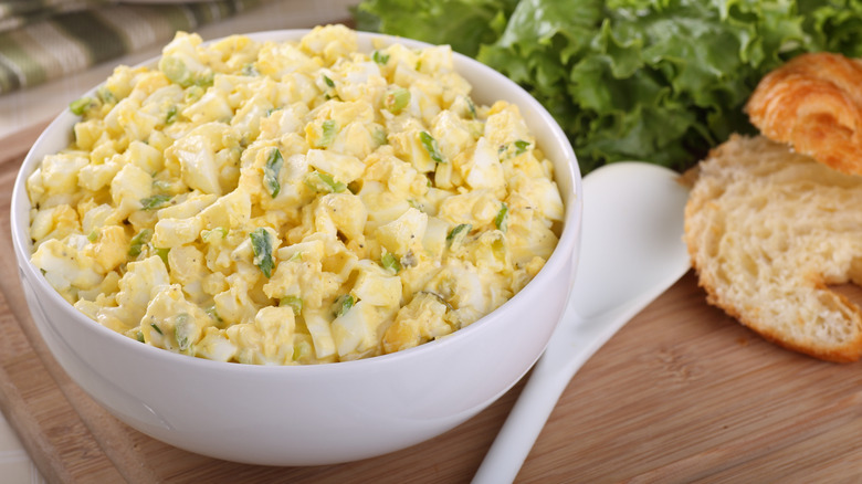 a ceramic bowl of egg salad on a cutting board beside a spoon, lettuce, and a sliced croissant