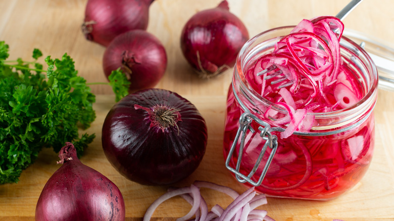 a fork scooping pickled onions from a jar sitting on a wooden table beside red onions and fresh herbs