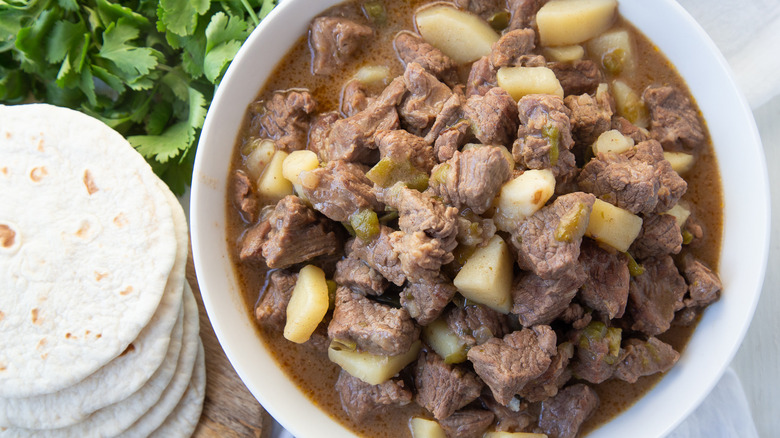 Bowl of carne guisada next to stack of flour tortillas and cilantro