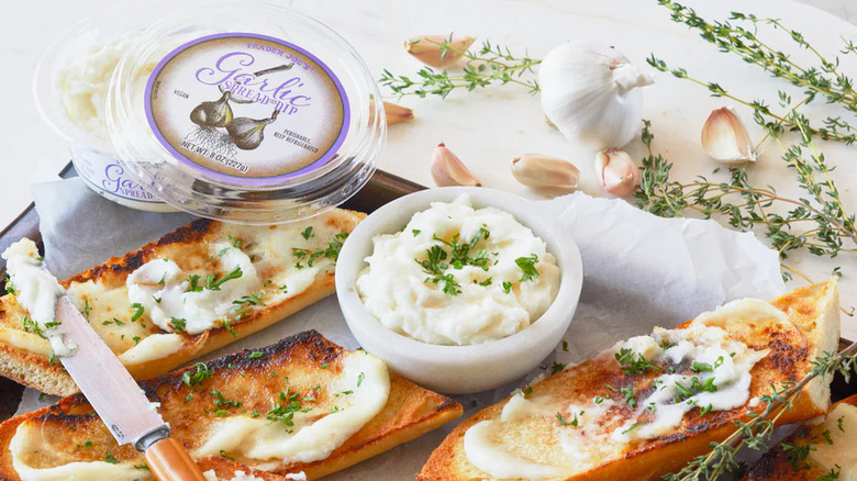 A container of Trader Joe's Garlic Spread Dip next to toasted bread and cloves of garlic