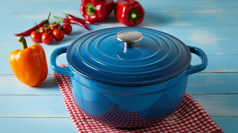 A bright blue Dutch oven on a blue wooden table surrounded by bell peppers, tomatoes, and chilis