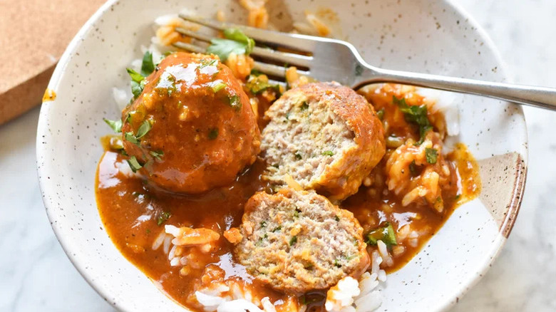 two meatballs, one sliced in half, coated in curry sauce sit in a bowl with rice and a fork.