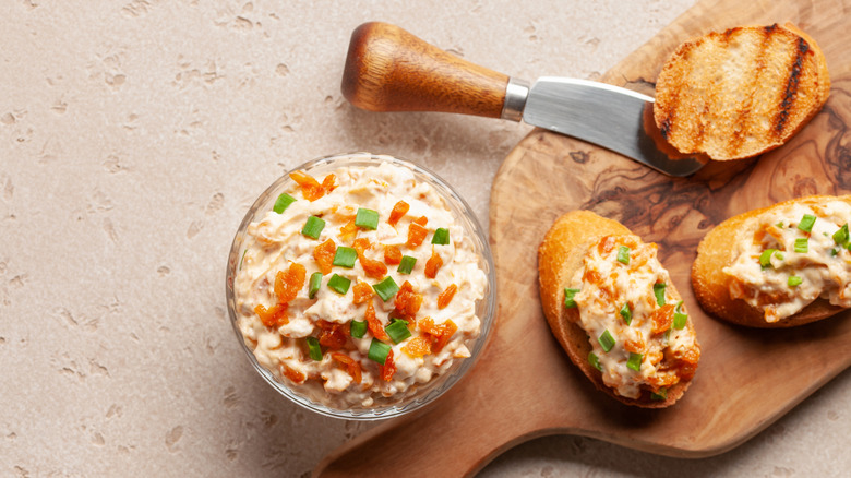 A small glass bowl of French onion dip next to three slices of toast, two with onion dip on cutting board.