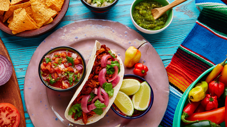 A plate with a taco, limes, and a bowl of salsa surrounded by bowls of salsa, peppers, and chips