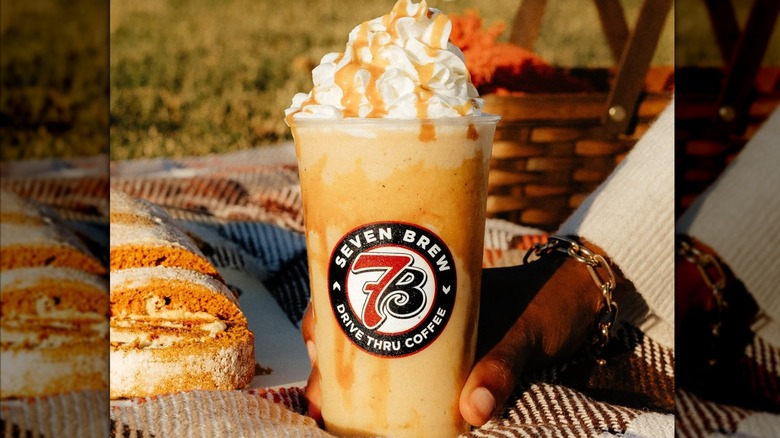 A 7 Brew pumpkin roll iced latte topped with whipped cream and drizzle being held on a picnic blanket next to a pumpkin Swiss roll in the sunshine
