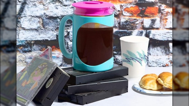 A turquoise and pink Primula Burke Cold Brew Coffee Maker on a kitchen counter next to croissants and a cup