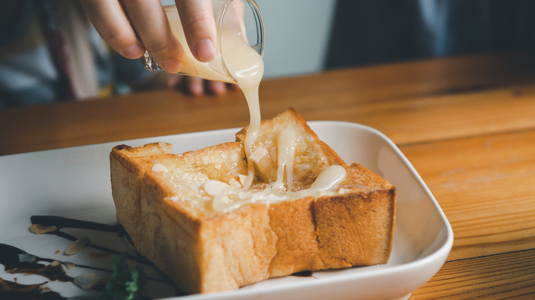 A hand pouring a drizzle of sweetened condensed milk from a small glass over a thick piece of square French toast