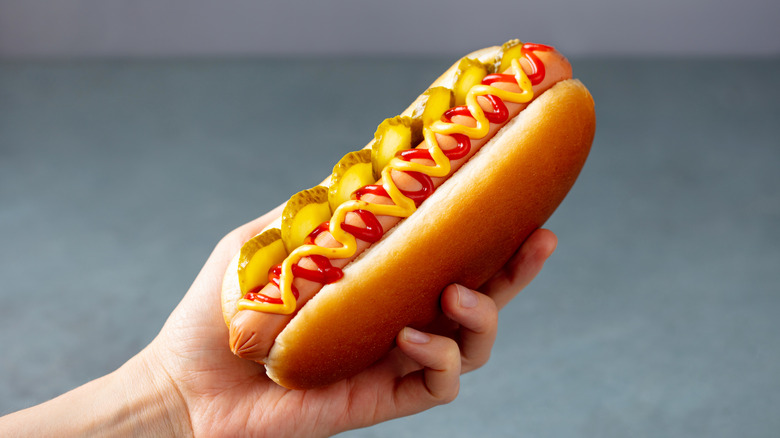 Hand holding a hot dog in a traditional bun with ketchup, mustard, and pickles