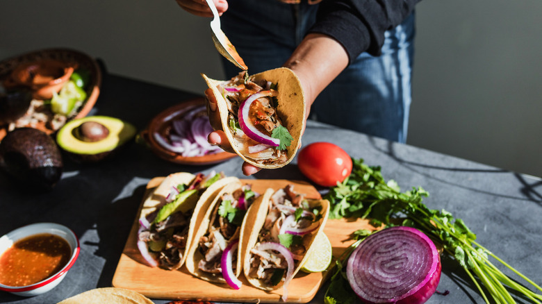 A woman holds a taco in one hand while drizzling sauce over its filling with a spoon, while three more tacos sit on a wooden board on a table in front of her, along with various taco ingredients including avocados and red onion