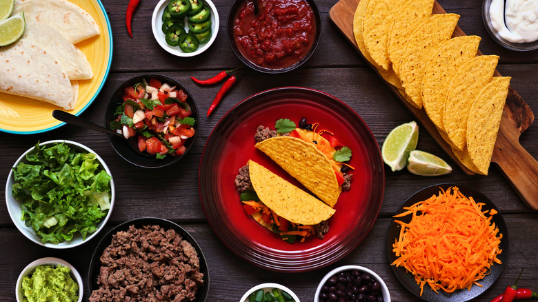 A table containing numerous plates and bowls of tacos and taco ingredients, including tortilla shells, shredded cheese, salsa, salad, cooked ground meat, lime wedges, and guacamole