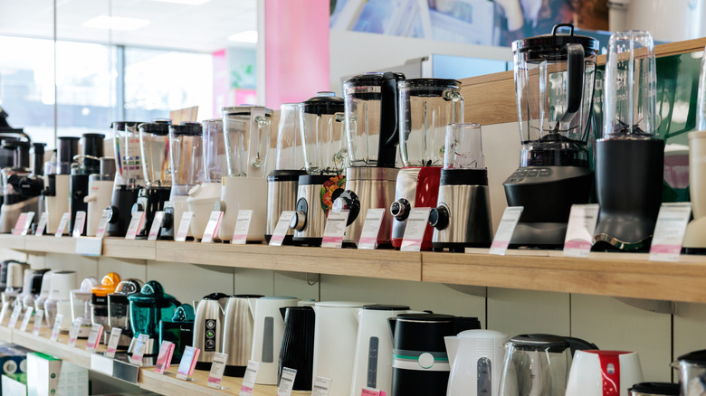 A small kitchen appliance store aisle with a shelf stocked full of blenders, a shelf of electric kettles are below them