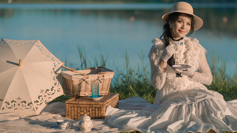 Woman in a Victorian era dress drinking tea beside a picnic basket and umbrella, with a lake in the background