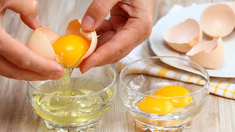 Hands separating egg whites from egg yolks into different bowls with egg shells on a plate in the background