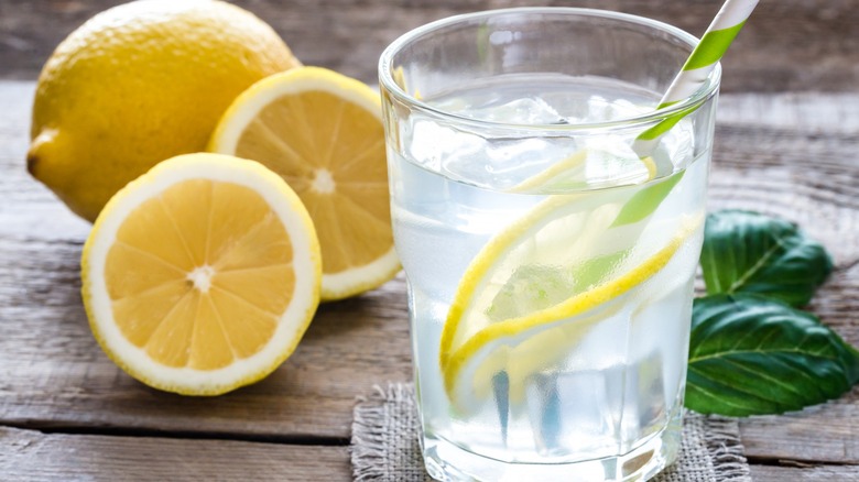 Glass of lemon ice water next to whole and sliced lemons